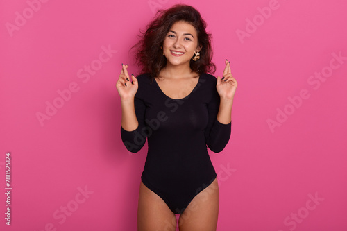 Indoor shot of pleasant looking brunette woman dressed black combidress  crosses fingers and has great hope  poses in pink studio  attractive female wishes fortune. Appealing girl with charming smile.