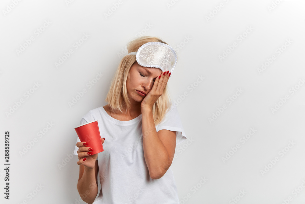 Tired sleepy young woman touching face with palm, has sad expression, closes eyes, holding disposable cup of drink. woman has problem with sleeping , isolated white background, studio shot Tired sleepy young woman touching face with palm, has sad expression, closes eyes, holding disposable cup of drink. woman has problem with sleeping , isolated white background, studio shot