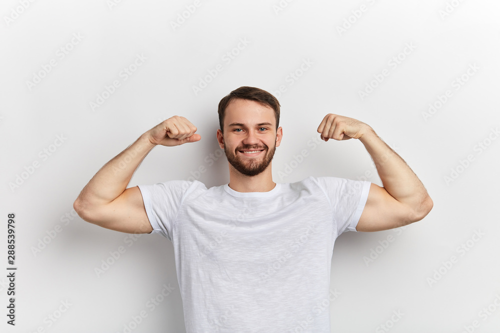 young handsome positive man wearing white t-shirt, holding his arm up to show his strength, biceps, strong arms, isolate white background. sport. hobby, interest, lifestyle, fitness, bodybuilding young handsome positive man wearing white t-shirt, holding his arm up to show his strength, biceps, strong arms, isolate white background. sport. hobby, interest, lifestyle, fitness, bodybuilding