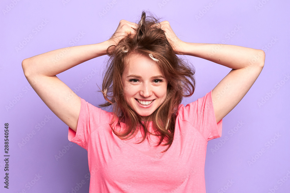 Photo of cheerful pretty girl smiling playfully, flirting with boy she likes at pyjamas pary, messing up her hair with hands after pillow fight, looking in camera, posing over purple wall. Photo of cheerful pretty girl smiling playfully, flirting with boy she likes at pyjamas pary, messing up her hair with hands after pillow fight, looking in camera, posing over purple wall.