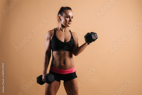 Portrait of muscular young woman with perfect athletic body in sports black bra posing with dumbbells. Shooting in professional studio on light pink red isolated background © dikushin
