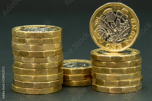 Three stacks of British one pound coins with one coin standing facing front on one of the stacks - all on a black background