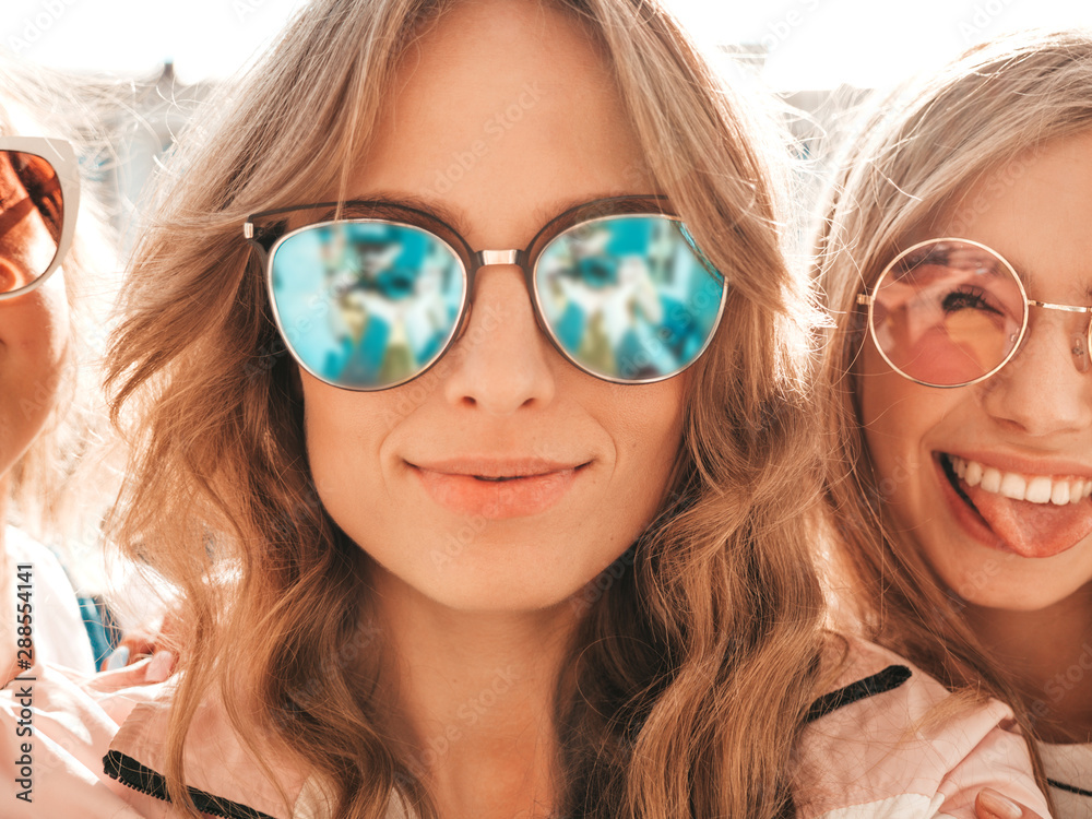 Three young smiling hipster women in summer clothes.Girls taking selfie self portrait photos on smartphone.Models posing in the street.Female showing positive face emotions in sunglasses