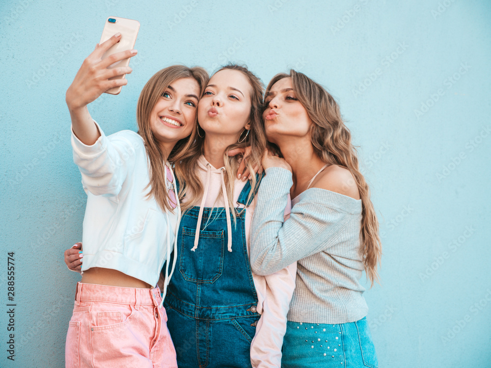 Three young smiling hipster women in summer clothes.Girls taking selfie self portrait photos on smartphone.Models posing in the street near wall.Female showing positive face emotions Three young smiling hipster women in summer clothes.Girls taking selfie self portrait photos on smartphone.Models posing in the street near wall.Female showing positive face emotions