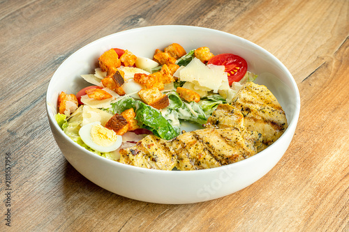 Caesar salad with fried chicken, croutons, parmesan, cherry tomatoes and white sauce in a white bowl on a wooden background. Restaurant serving. traditional lunch