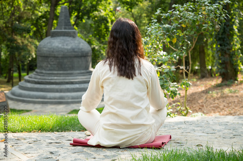 Woman sitting in yoga pose view from the back. Meditation on the background of the temple and nature. Engage in a retreat center.