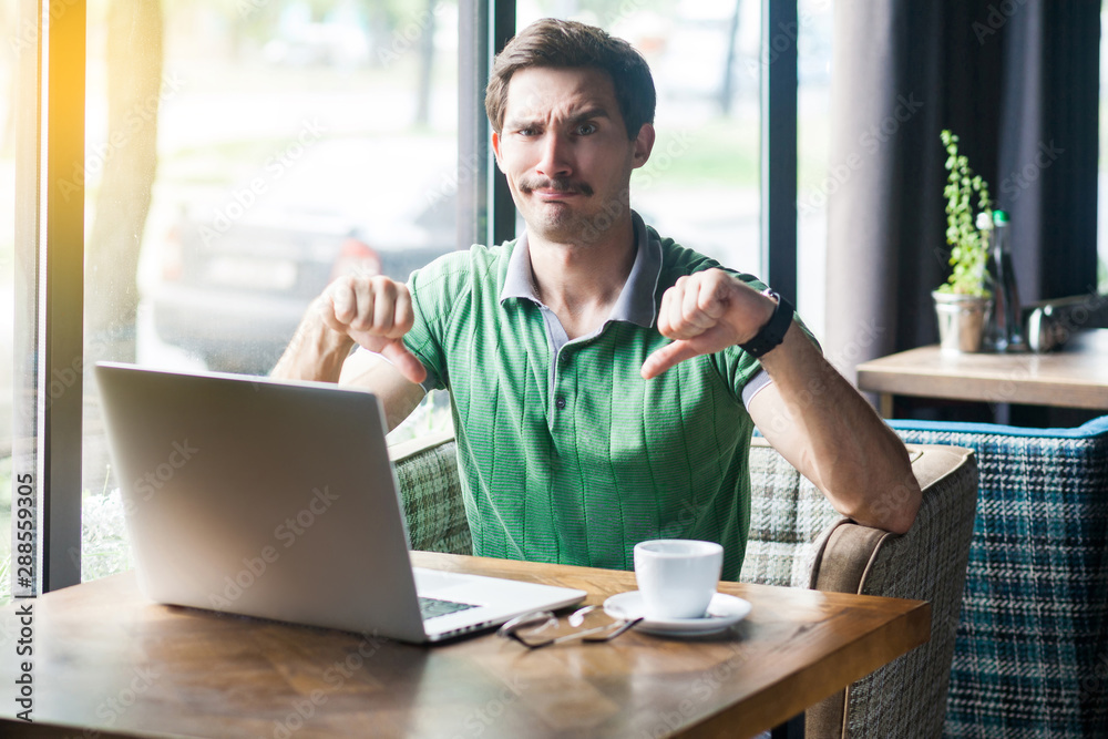 Dislike! Young dissatisfied businessman in green t-shirt sitting, working on laptop, looking at camera and showing thumbs down. business and freelancing concept. indoor shot near big window at daytime Dislike! Young dissatisfied businessman in green t-shirt sitting, working on laptop, looking at camera and showing thumbs down. business and freelancing concept. indoor shot near big window at daytime
