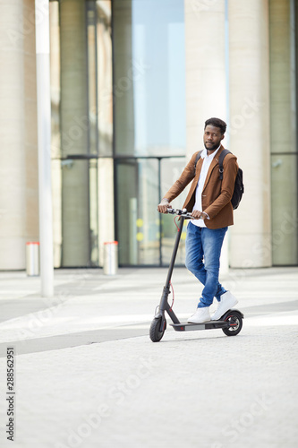 Full length portrait of trendy African man riding electric scooter and looking at camera while commuting in city street, copy space