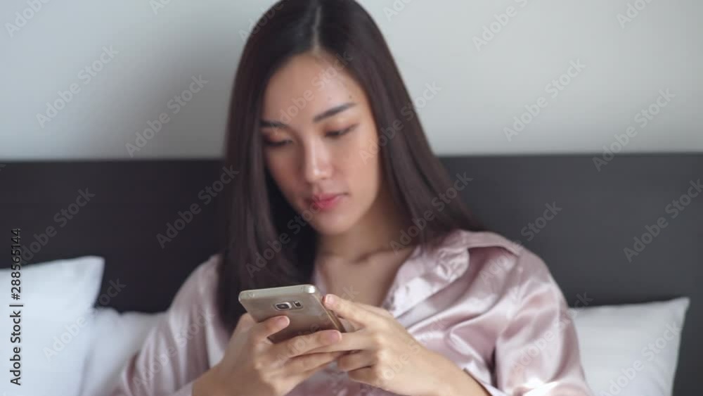 cheery young Asian woman using mobile phone while sitting on her bed. Focus on smartphone. low light cheery young Asian woman using mobile phone while sitting on her bed. Focus on smartphone. low light