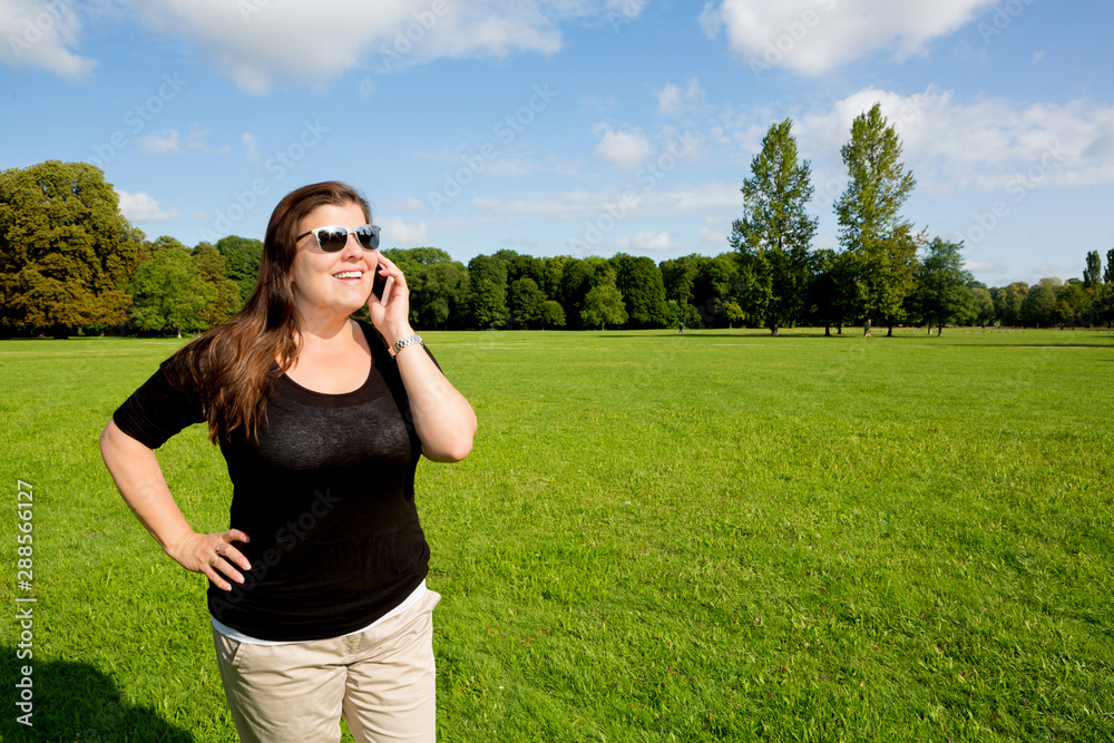 Woman speaks on the phone in a public green park during a summer day. She is receiving some good news.