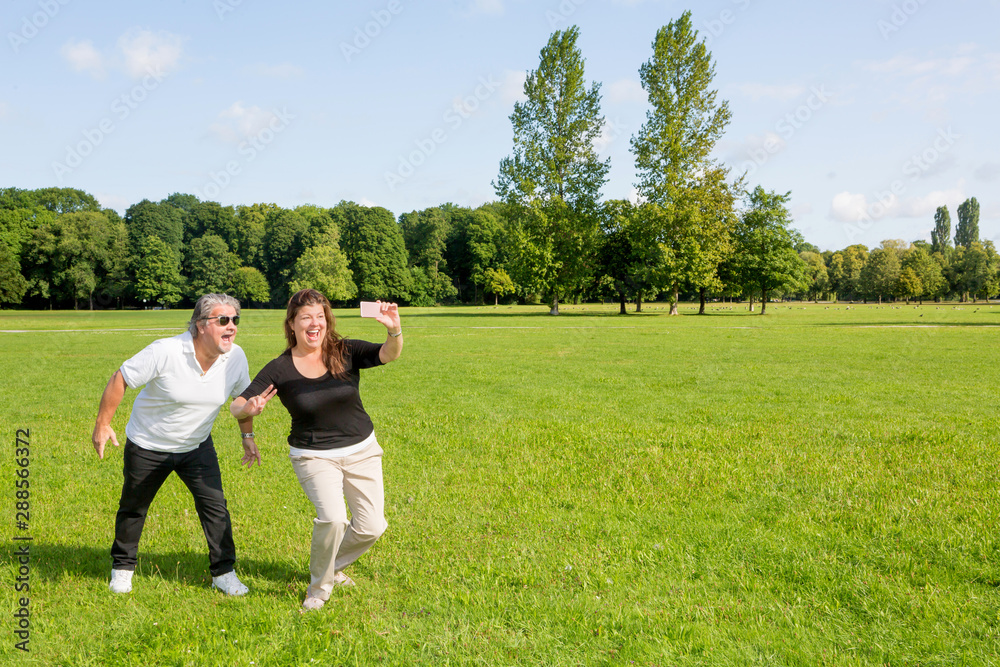 A man and a woman cheerfully take a selfie together as celebration. They are outside in a public park during a sunny summer day. A man and a woman cheerfully take a selfie together as celebration. They are outside in a public park during a sunny summer day.