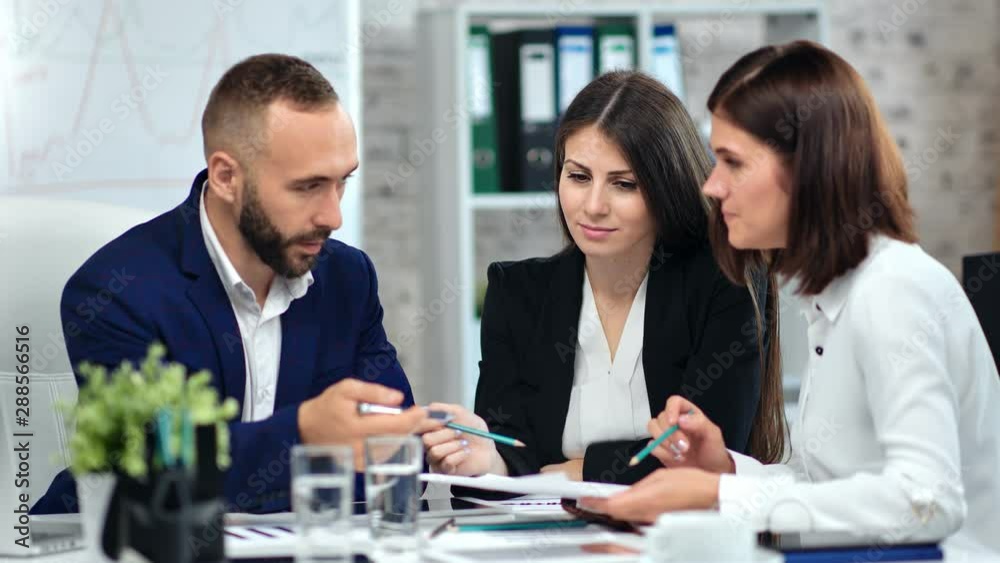 Male trendy boss in suit discussing project with two female employee at modern office interior Male trendy boss in suit discussing project with two female employee at modern office interior