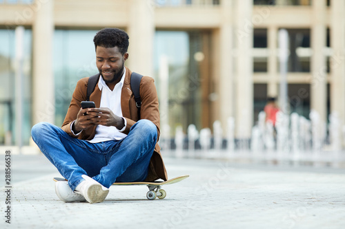 Full length portrait of contemporary African-American man using smartphone and smiling while sitting cross legged on skateboard outdoors, copy space