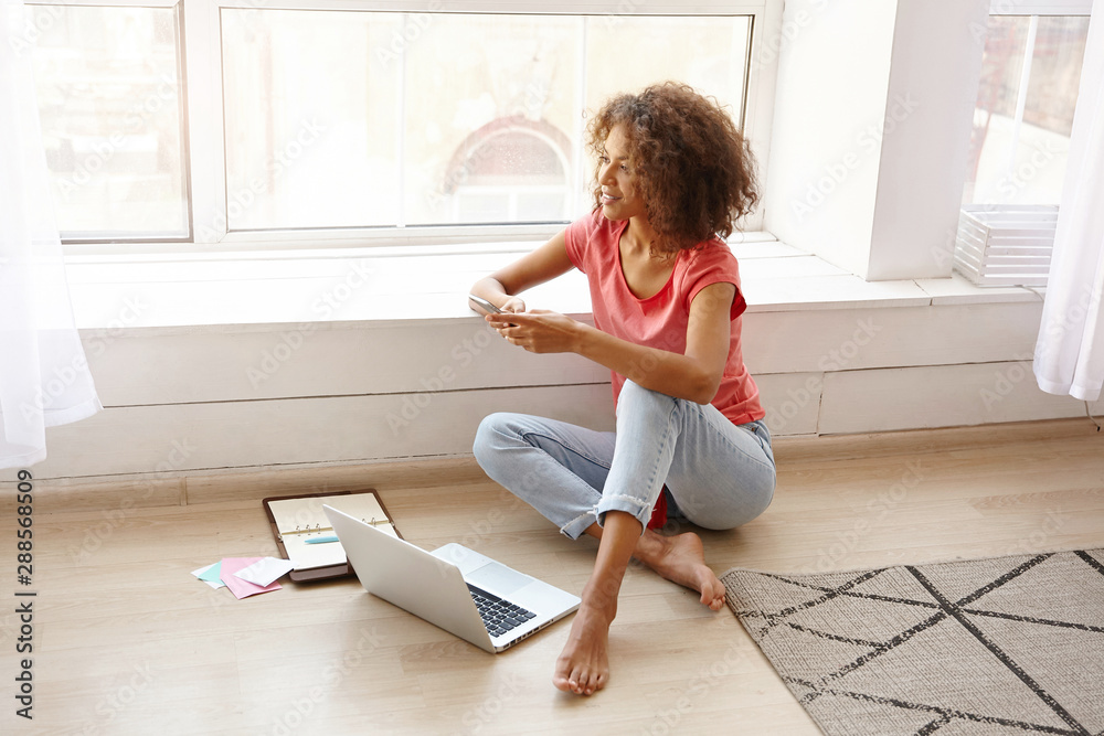 Indoor photo of attractive curly female with dark skin leaning on windowsill and looking ahead, wearing pink t-shirt and blue jeans, holding mobile phone in hands Indoor photo of attractive curly female with dark skin leaning on windowsill and looking ahead, wearing pink t-shirt and blue jeans, holding mobile phone in hands