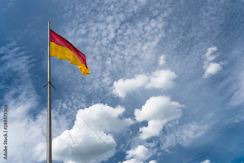 Flag in red at the top and yellow at the bottom, hanging on the mast, waving in the wind. In the background a blue sky with white clouds.