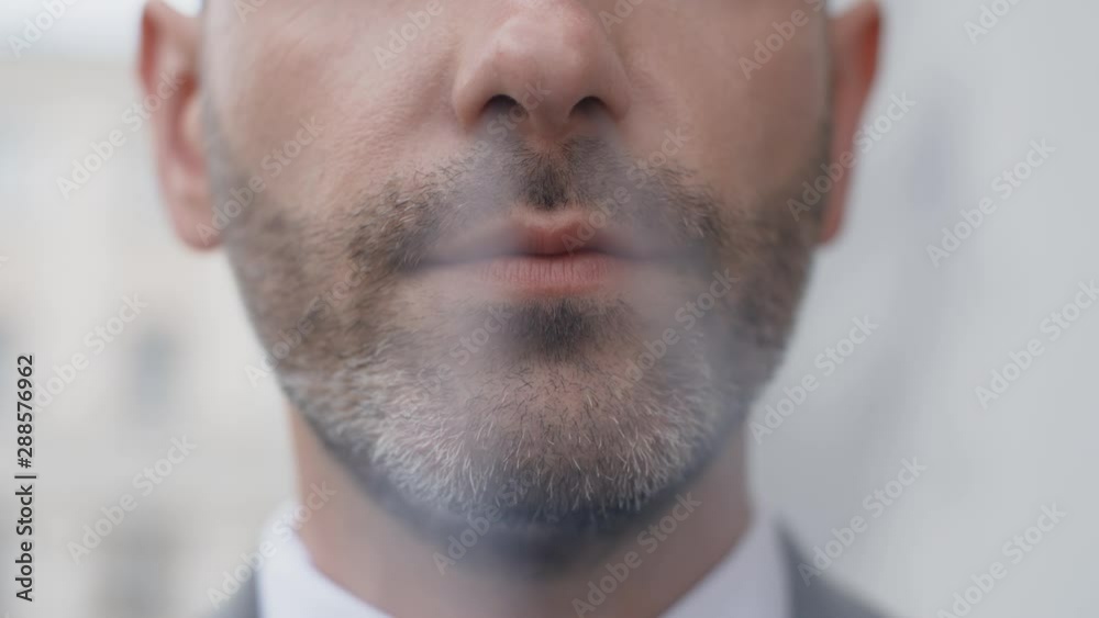 Extreme close face of attractive middle aged bearded man in business suit smoking cigarette in front of camera outside. Portrait of handsome male businessman standing outdoors