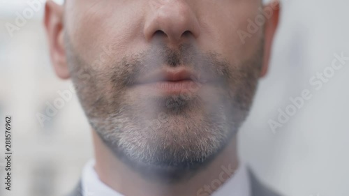 Extreme close face of attractive middle aged bearded man in business suit smoking cigarette in front of camera outside. Portrait of handsome male businessman standing outdoors photo