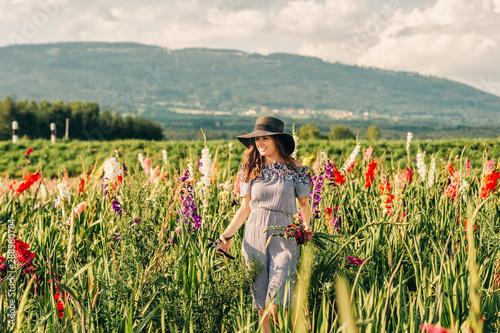 Gorgeous young woman picking flowers in a field, wearing summer dress, black straw hat