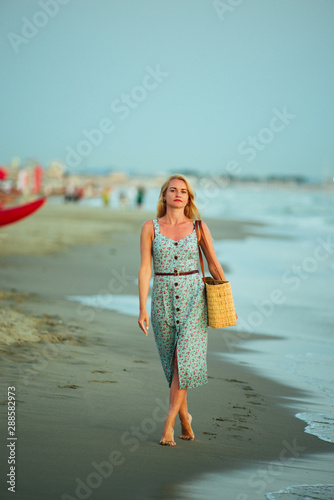 Blonde caucasian woman with an elegant fashionable dress and her wooden bag posing in a portrait session near the sea at sunset