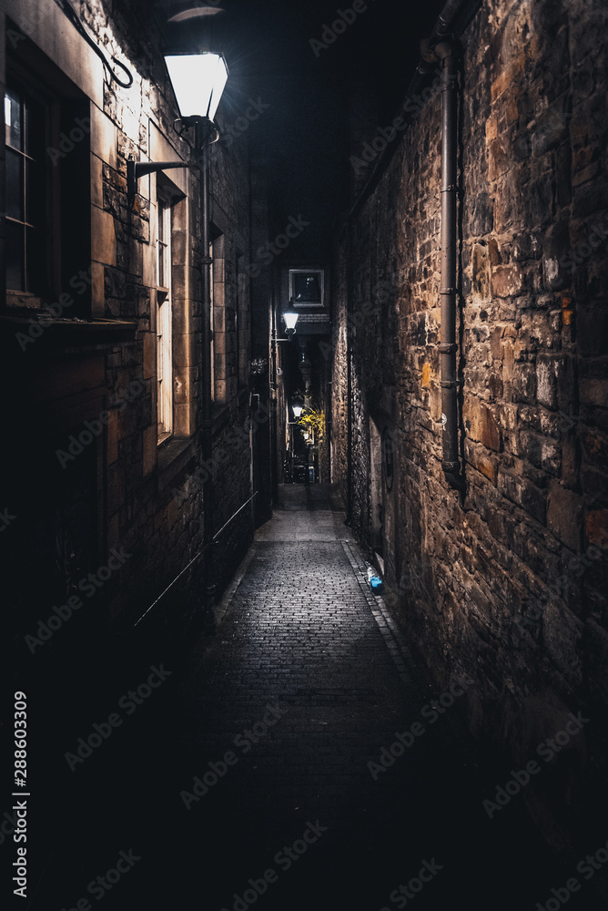 Fototapeta A dark creepy narrow European alley at night, surrounded by bricks and cobblestone. Illuminated only with some street lamps. Concept of scared or being alone and frightened