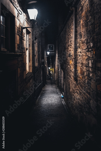 A dark creepy narrow European alley at night  surrounded by bricks and cobblestone. Illuminated only with some street lamps. Concept of scared or being alone and frightened