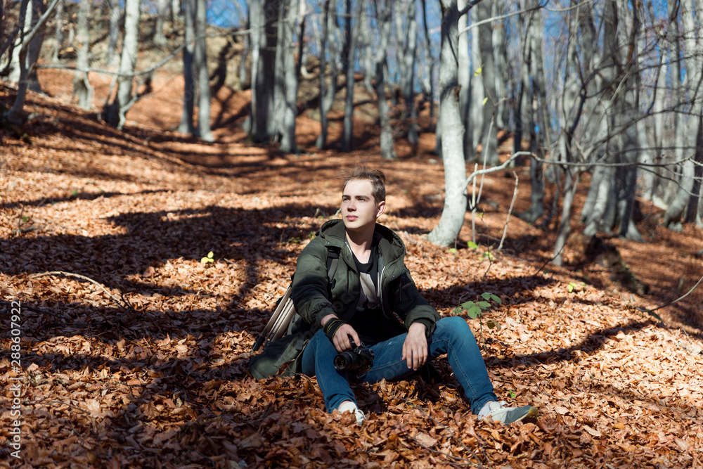 A young attractive guy is sitting in autumn beech forest on yellow foliage. In his hands he holds a camera.