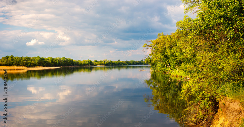 The calm river with the cloudy stormy sky and the green forest, lit by sunlight