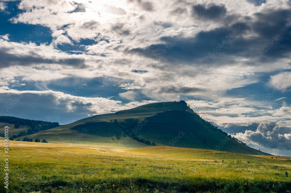 Grass hills covered with trees in steppe under spectacular clouds sky during sunset at Khakassia, Siberia, Russia.