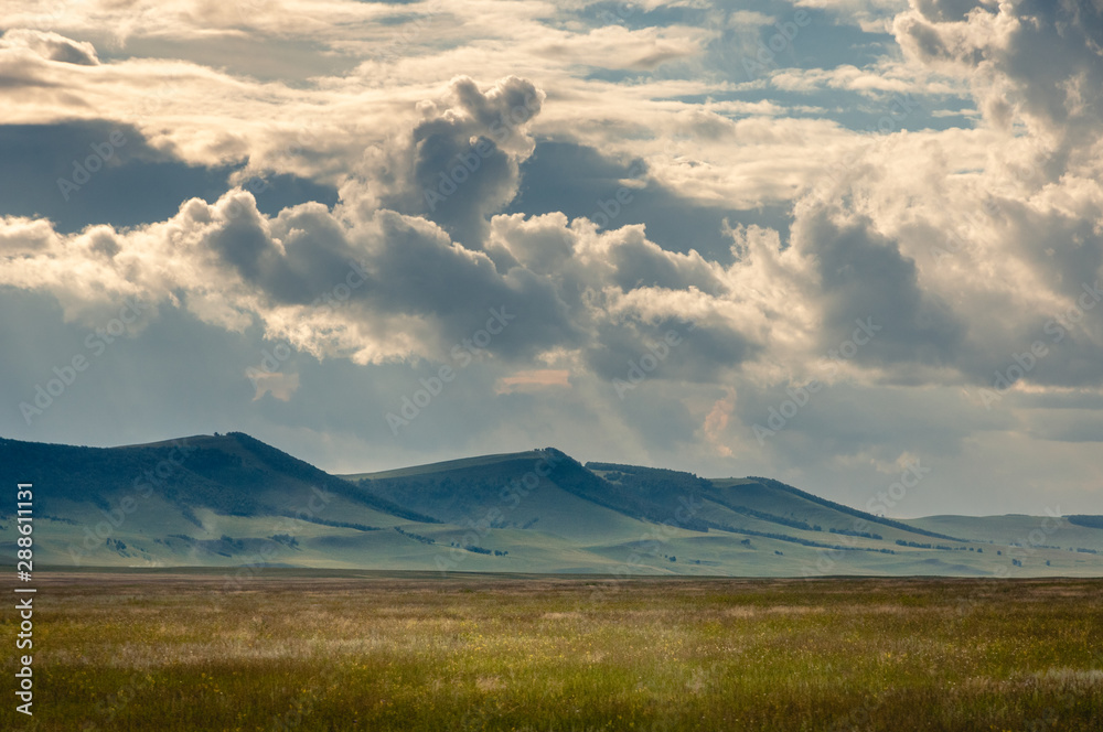 Grass hills on horizon in steppe under heavy clouds sky during sunset with sun light beams at Khakassia, Siberia, Russia.