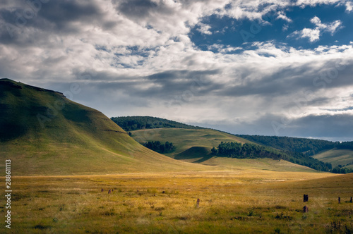 Grass hills covered with trees in steppe under spectacular clouds sky during sunset at Khakassia, Siberia, Russia.