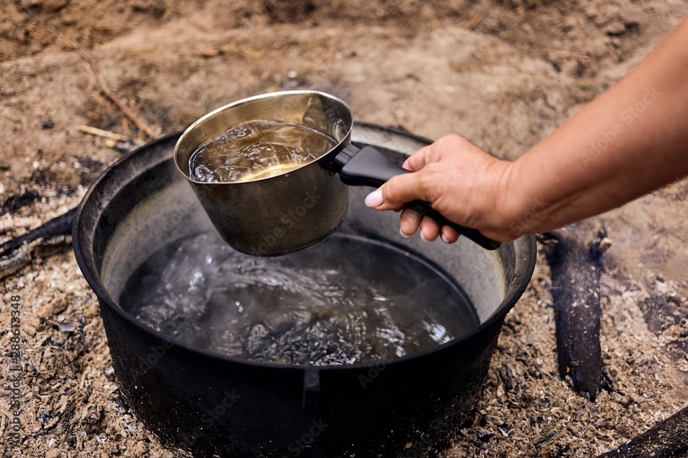 Boiling water in cauldron. Woman's hand taking water in cauldron with a kettle for cooking in the forest. Boiling water in cauldron. Woman's hand taking water in cauldron with a kettle for cooking in the forest.