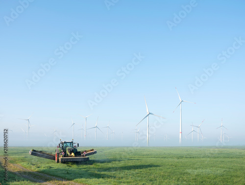 farmer mows grass near wind turbine farm in oastfriesland on misty summer morning