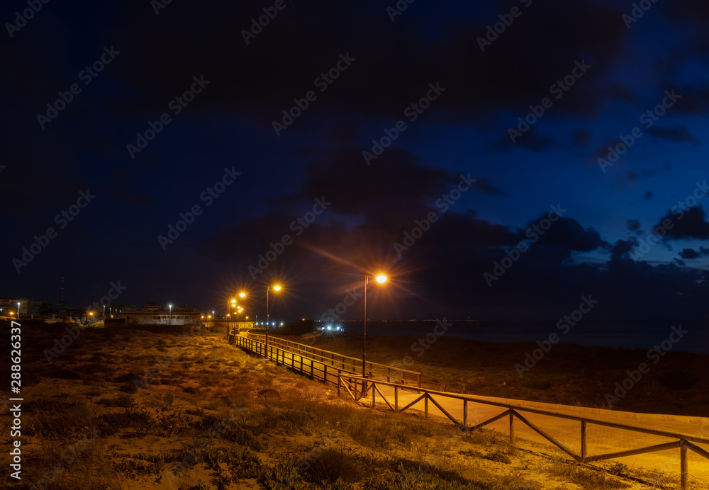 Blue hour with lanterns on the wooden path on the beach of Torrevieja in the early morning at dusk. The wooden bridge leads along the beach. In the background the Mediterranean sea and the coastline.