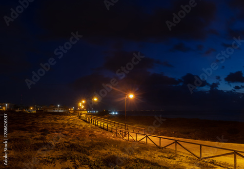 Blue hour with lanterns on the wooden path on the beach of Torrevieja in the early morning at dusk. The wooden bridge leads along the beach. In the background the Mediterranean sea and the coastline.