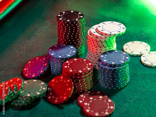 Close-up top view of colorful chips piles, some of them laying nearby on green cover of playing table, under green, red neon light. Black background.