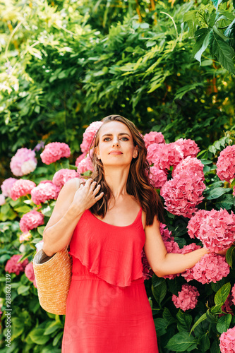 Outdoor portrait of beautiful woman posing in pink hydrangea flowers, wearing red dress