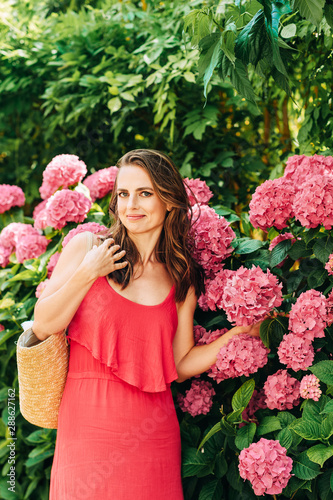 Outdoor portrait of beautiful woman posing in pink hydrangea flowers, wearing red dress