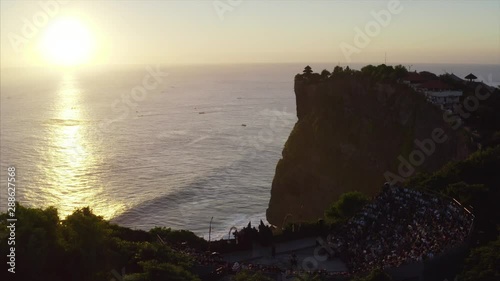 Aerial static view of outdoor round stage with many spectators and dance Kecak performance. Bright sunset and fishing boats on ocean surface. Uluwatu temple. Bali, Indonesia photo