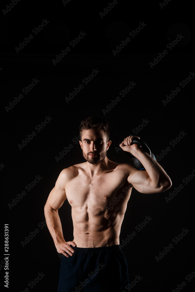 Young bearded man with a kettlebell in his hands on a black background. bare-chested athlete