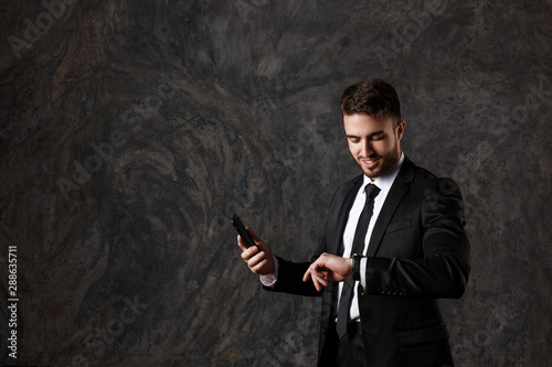 a young bearded man looks at his watch and holds a mobile phone in his hands. successful businessman in a suit watches time on a wristwatch