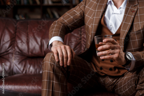 young successful man in a brown kosmtyum drinks whiskey. bearded businessman sitting in a library on a luxurious leather sofa and drinking cognac