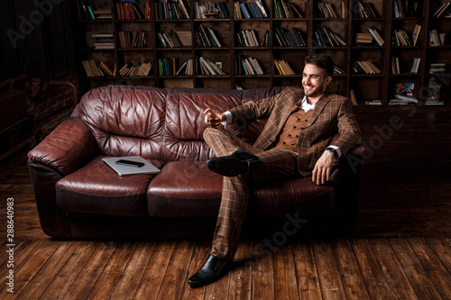 young successful man in a brown kosmtyum drinks whiskey. bearded businessman sitting in a library on a luxurious leather sofa and drinking cognac photo