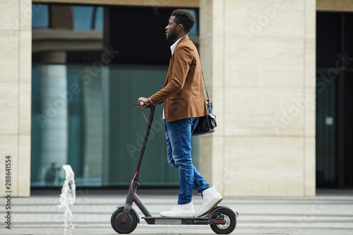 Side view full length of modern African-American businessman riding electric scooter while commuting to work in city, copy space