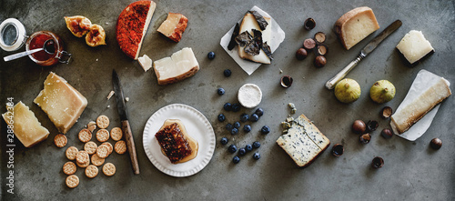 Wine snack variety. Flat-lay of cheeses, honey, fresh fruit, nuts and crackers over rough grey concrete background, top view, wide composition. Gathering, party, holiday food concept photo