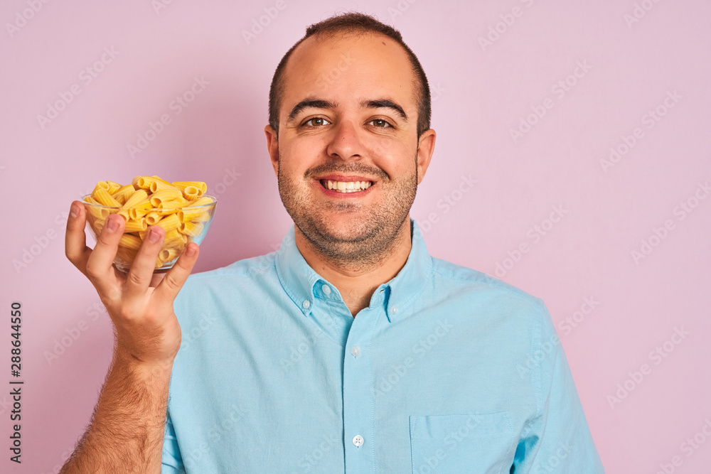Young man holding bowl with macaroni pasta standing over isolated pink background with a happy face standing and smiling with a confident smile showing teeth Young man holding bowl with macaroni pasta standing over isolated pink background with a happy face standing and smiling with a confident smile showing teeth