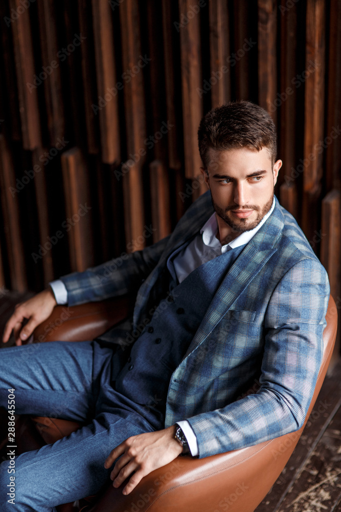 Young successful businessman in a blue suit in a cage is sitting in a leather chair. young man with a beard smiling. Young successful businessman in a blue suit in a cage is sitting in a leather chair. young man with a beard smiling.