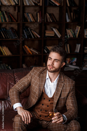 young successful man in a brown kosmtyum drinks whiskey. bearded businessman sitting in a library on a luxurious leather sofa and drinking cognac