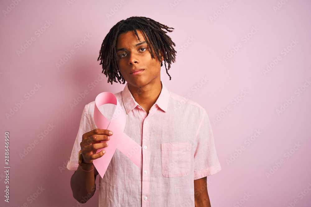 Afro american man with dreadlocks holding cancer ribbon over isolated pink background with a confident expression on smart face thinking serious Afro american man with dreadlocks holding cancer ribbon over isolated pink background with a confident expression on smart face thinking serious