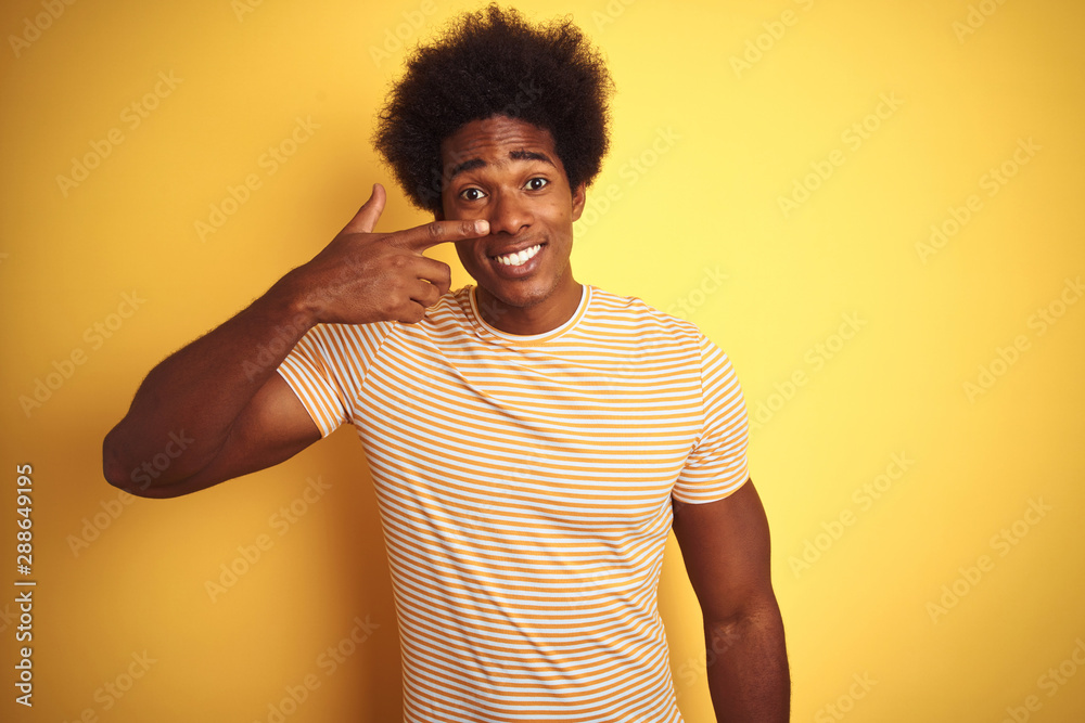 American man with afro hair wearing striped t-shirt standing over isolated yellow background Pointing with hand finger to face and nose, smiling cheerful. Beauty concept American man with afro hair wearing striped t-shirt standing over isolated yellow background Pointing with hand finger to face and nose, smiling cheerful. Beauty concept