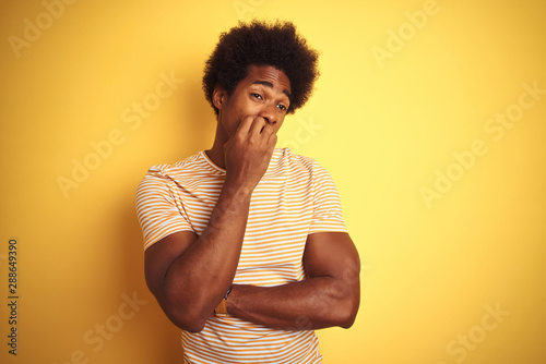 American man with afro hair wearing striped t-shirt standing over isolated yellow background looking stressed and nervous with hands on mouth biting nails. Anxiety problem.